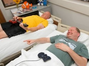 Dois senhores em camas separadas jogando videogame e tocando guitarra